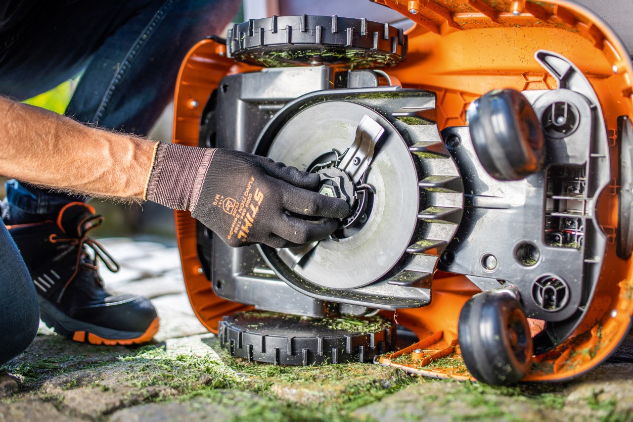 A STIHL iMOW® 422 robot mower tipped up on its side while a person wearing gloves inspects the blade